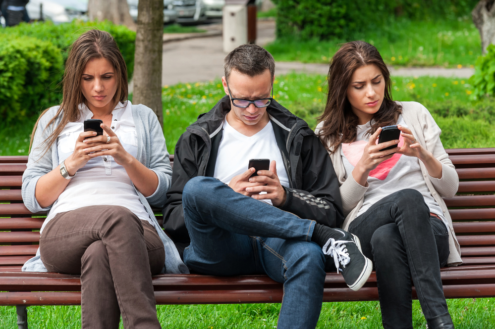 Three adolescents on a park bench. They are ignoring each other while on their phones.