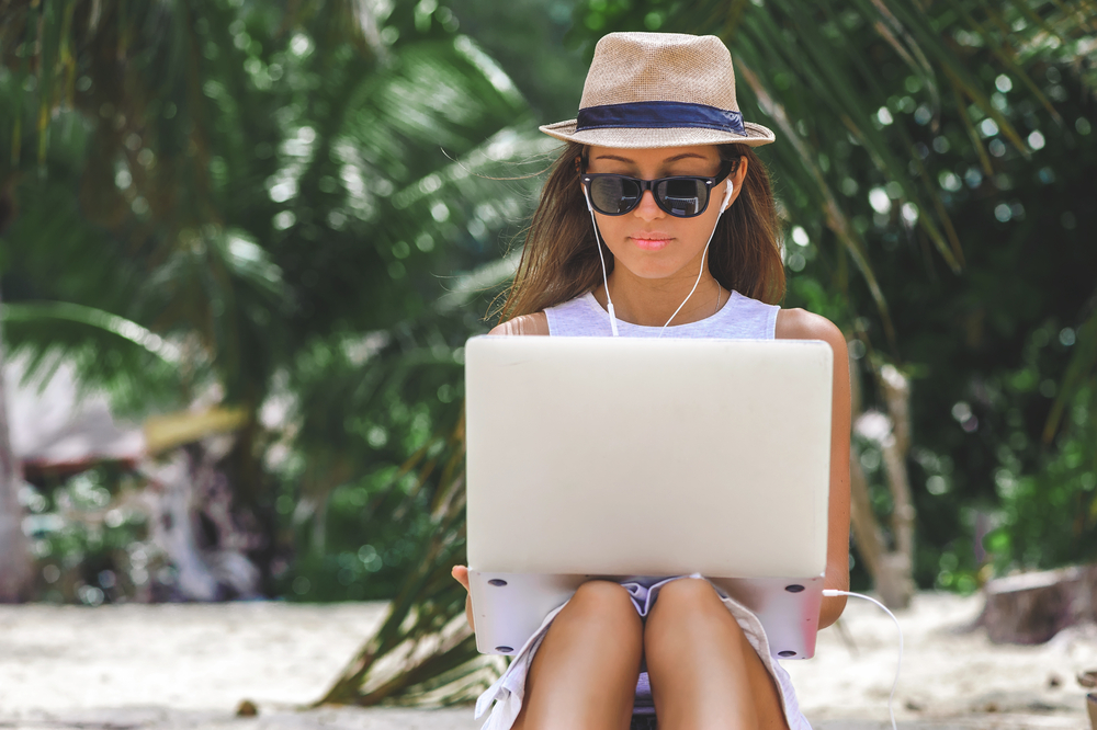 Young attractive woman in dress, sunglasses and hat, working in laptop on the beach, on palm trees background, Girl Freelancer working
