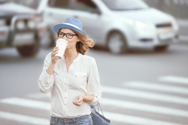 Happy young fashionable student drinking hot take away coffee and crossing a road against urban background.