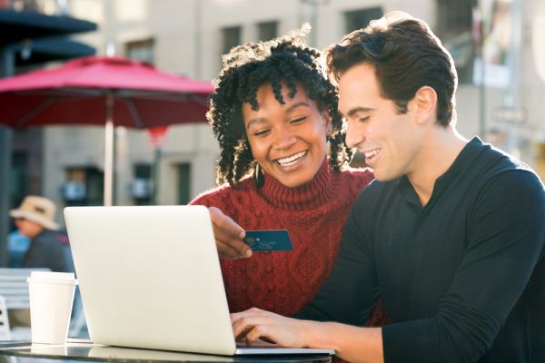 Beautiful young loving couple shopping online together while woman holding credit card and smiling. Portrait of happy couple paying bills online using laptop and credit card. Happy smiling couple with laptop and credit card buying online through ecommerce.