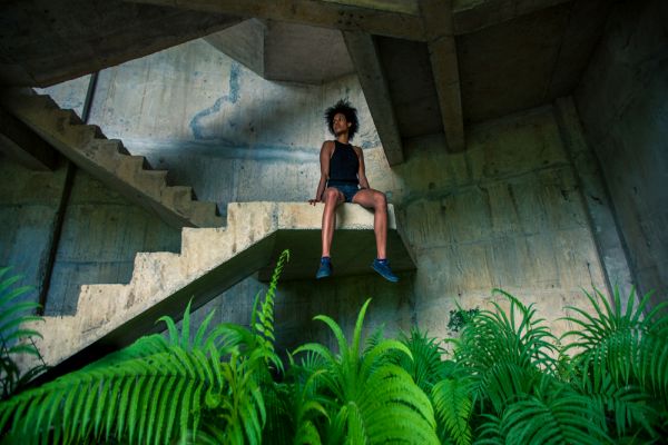 A woman sitting on the landing of a stairwell that is dangling over bright green ferns.
