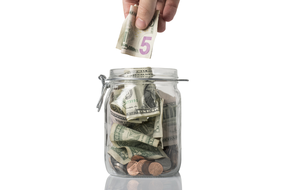 A tip or savings jar filled with American coins and bills and someone about to add a five dollar bill.