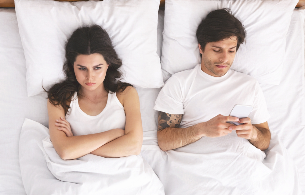 Problem in bed. Addicted man playing video game on cellphone, offended wife lying next to him, top view