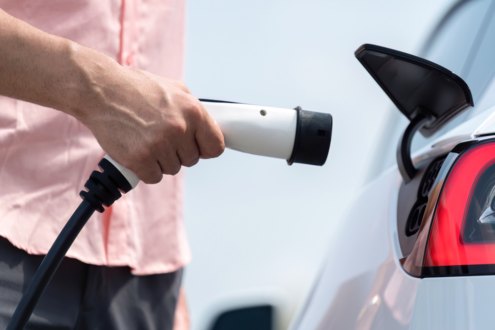 Hand insert EV charger plug into electric vehicle to recharge EV car battery from outdoor charging station. Fresh daylight environment with alternative clean and sustainable energy concept.