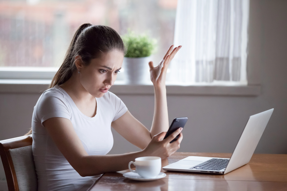 A woman looking at her phone with frustration, sitting in front of a laptop.