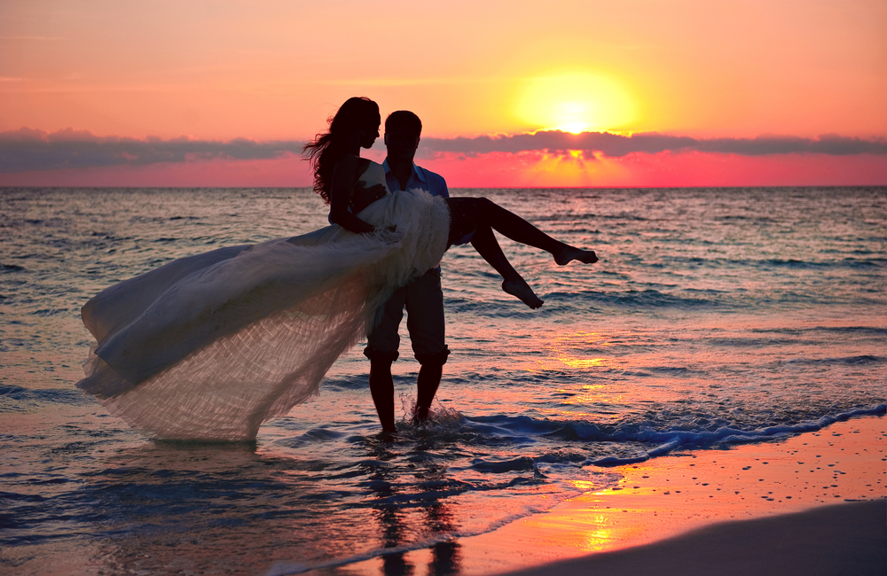 Silhouettes of a bride and groom in ankle-deep water at the beech during sunset.